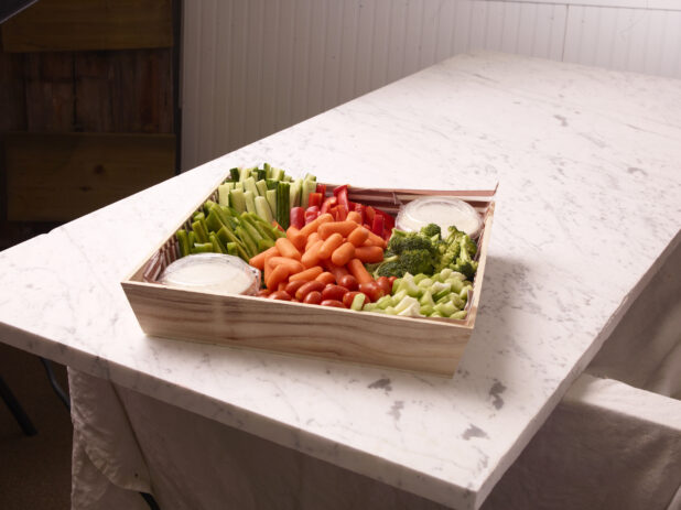 Wood catering tray with assorted vegetables and two creamy dips on a white marble countertop