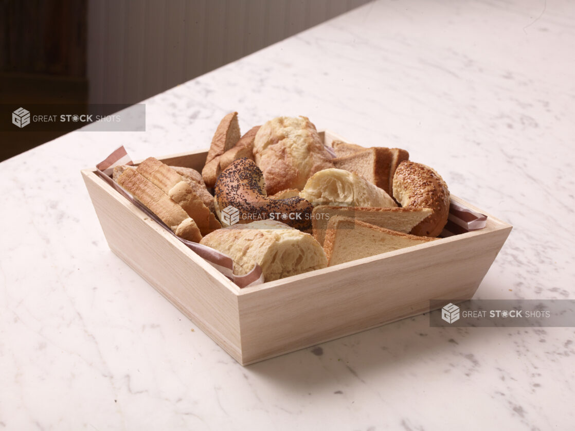 Wood catering tray filled with assorted breads, buns and bagels on a white marble countertop