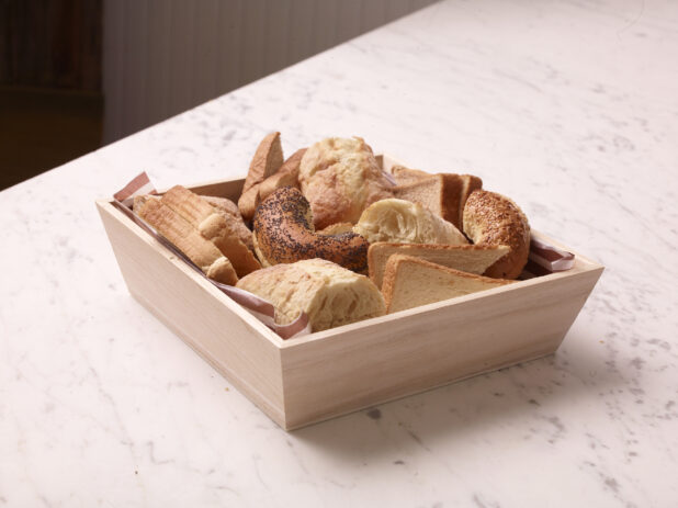 Wood catering tray filled with assorted breads, buns and bagels on a white marble countertop