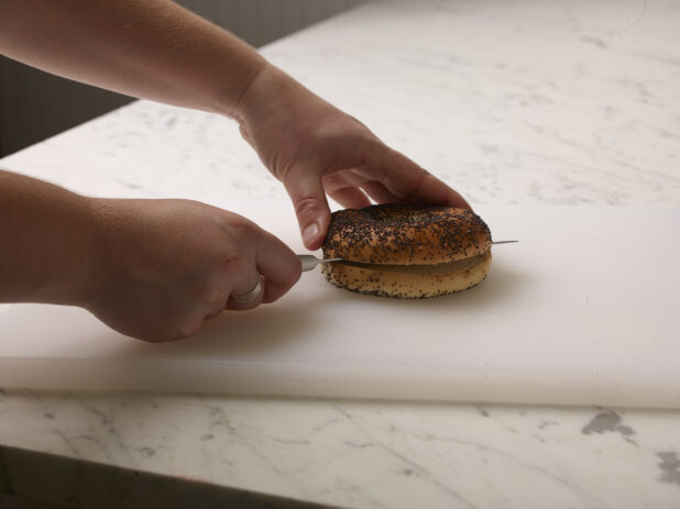 Woman cutting a poppyseed bagel in half on a white cutting board with a white marble background