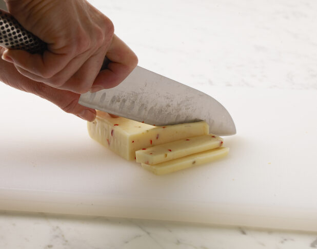 Woman cutting a brick of Monterey Jack cheese into slices on a white cutting board with a white marble background