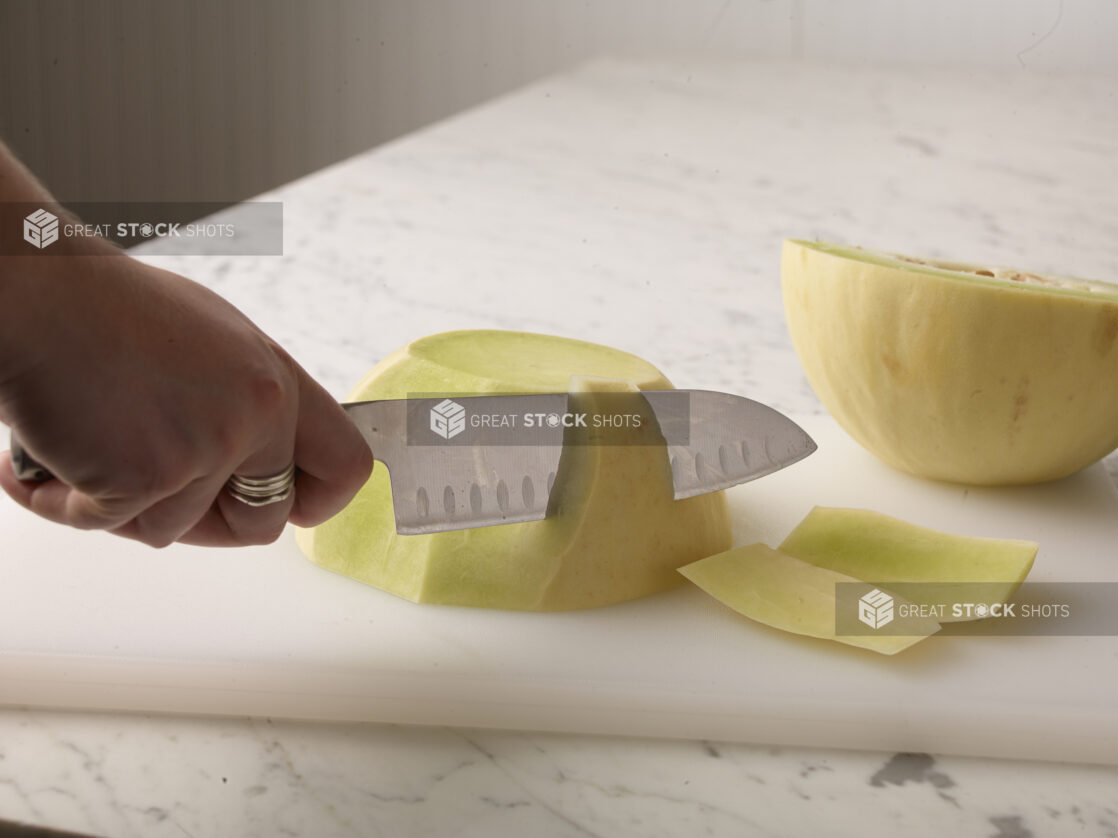 Hand cutting a honeydew melon with a kitchen knife on a white cutting board on a white marble background