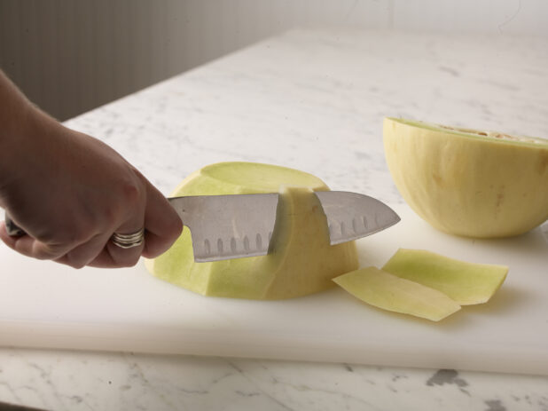 Hand cutting a honeydew melon with a kitchen knife on a white cutting board on a white marble background