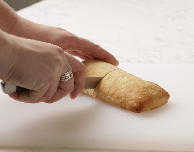 Woman cutting a white bun in half on a white cutting board with a white marble background