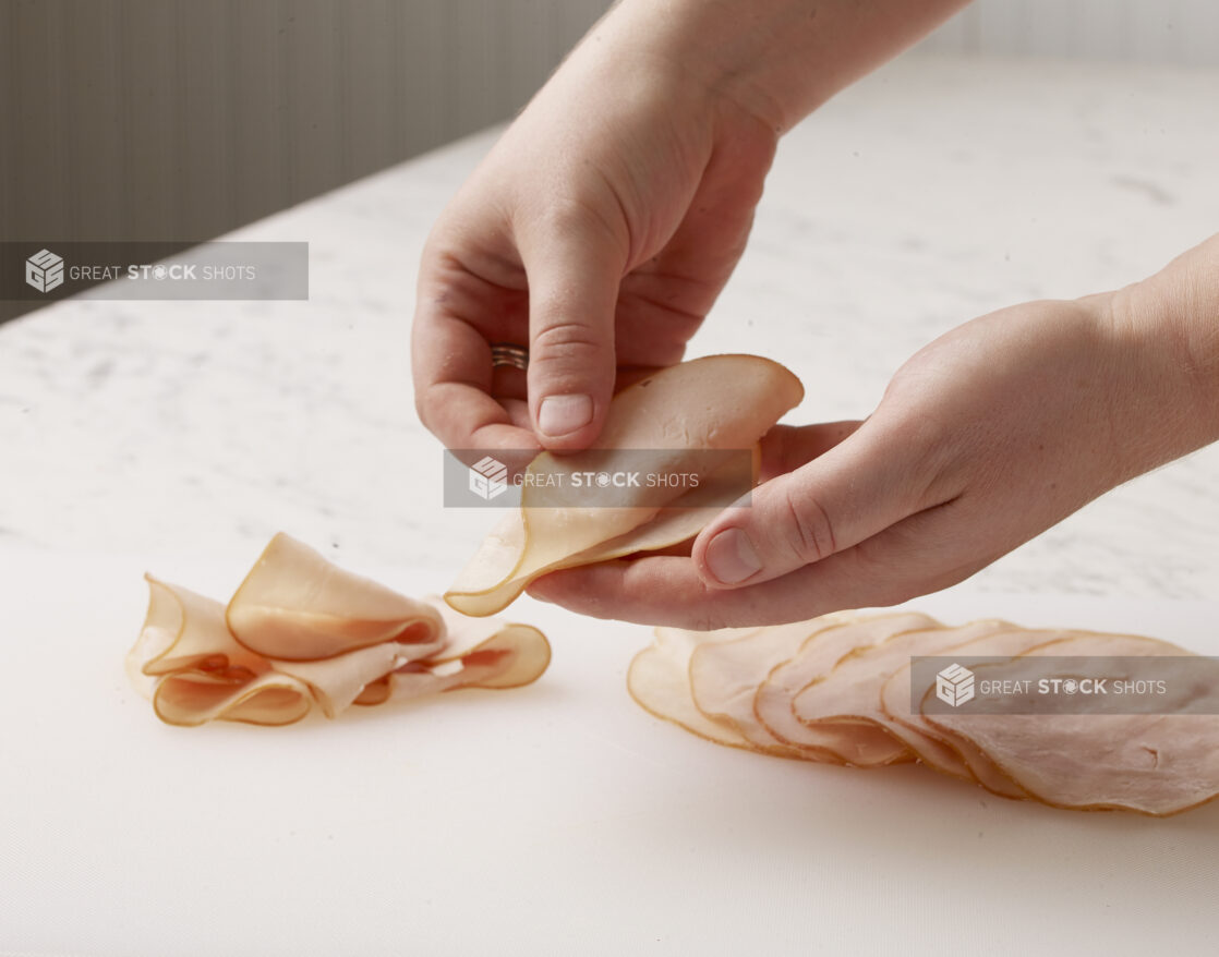 Hands folding sliced oven roasted turkey meat on a white cutting board with a white marble background