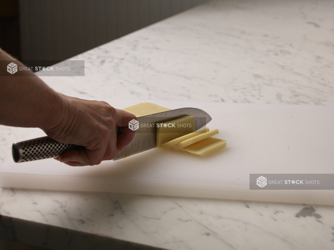 Woman cutting a brick of cheese into slices on a white cutting board with a white marble background