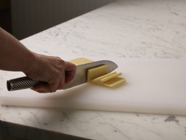 Woman cutting a brick of cheese into slices on a white cutting board with a white marble background