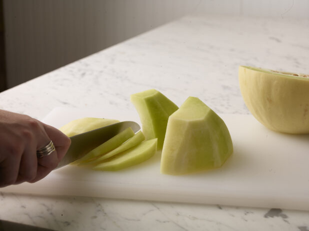 Hand using a kitchen knife to cut a honeydew melon on a white cutting board on a white marble background