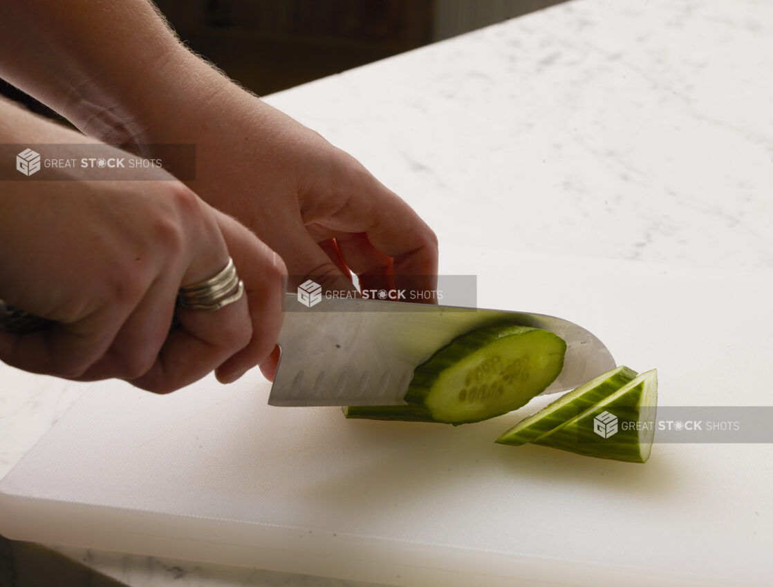 Woman slicing a cucumber diagonally on a white cutting board with a white marble background