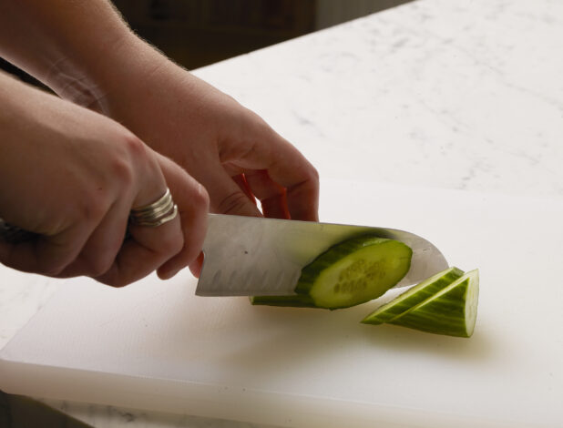 Woman slicing a cucumber diagonally on a white cutting board with a white marble background
