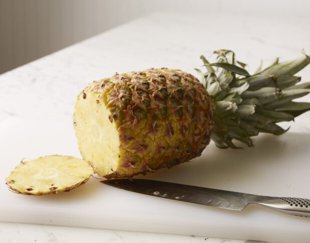 Whole pineapple with bottom end cut off on a white cutting board with a chefs knife on the board