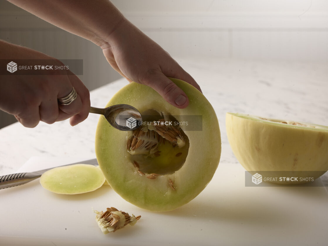 Hands holding half a honeydew melon cleaning out the seeds with the other half on a white cutting board