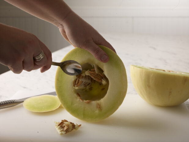 Hands holding half a honeydew melon cleaning out the seeds with the other half on a white cutting board