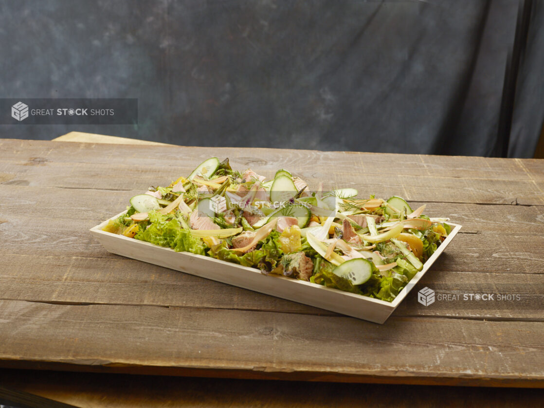 Wood catering tray of salmon salad with peaches, cucumber, shaved carrot and asparagus on mixed greens on a wood table