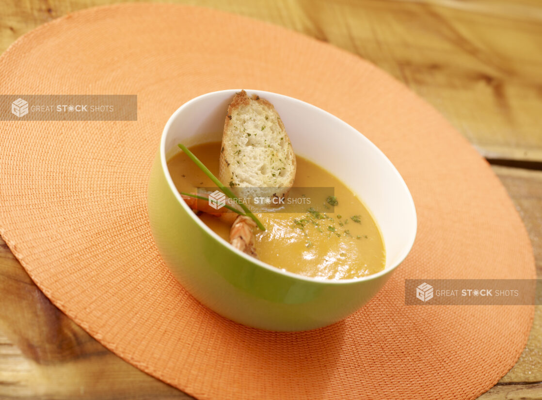 Bisque Soup in Green Bowl with Shrimp, Chives and Sliced Baguette on a Wooden Table with Bokeh Effect