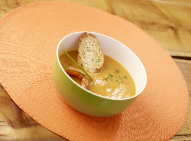 Bisque Soup in Green Bowl with Shrimp, Chives and Sliced Baguette on a Wooden Table with Bokeh Effect
