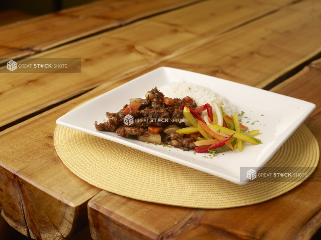 Jamaican Brown Stew with White Rice and Raw Vegetables on a Square White Dish on a Wooden Table with Bokeh Effect