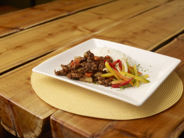 Jamaican Brown Stew with White Rice and Raw Vegetables on a Square White Dish on a Wooden Table with Bokeh Effect