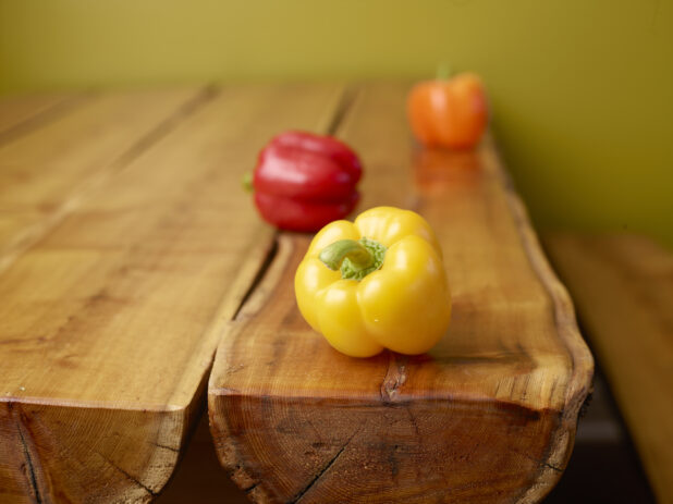 Whole Yellow, Red and Orange Bell Peppers on a Wooden Table with Green Background and Bokeh Effect