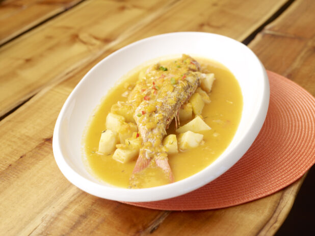 Whole Red Snapper Stew with Potatoes in a Large White Platter on an Orange Placemat on a Wooden Table