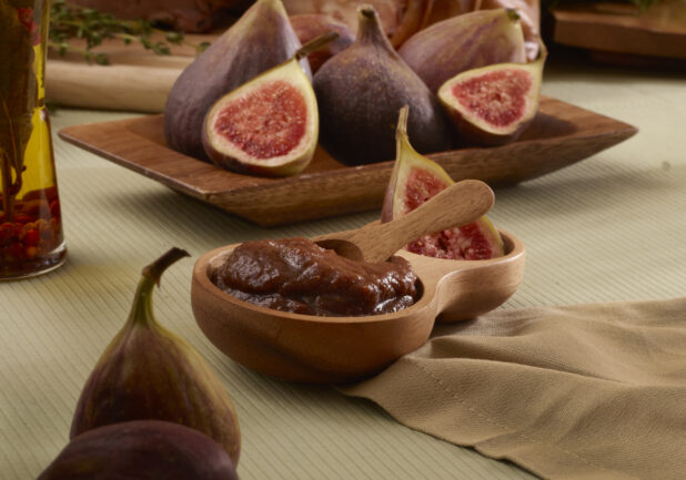 Wooden Bowl of Fig Jam with Fresh Whole and Cut Figs in Background on a Green Table Cloth Surface in an Indoor Setting
