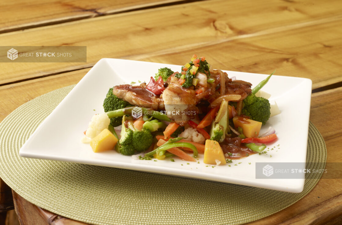 Fried White Fish with Broccoli and Sautéed Vegetables on a Square White Plate on a Green Placemat on a Wooden Table