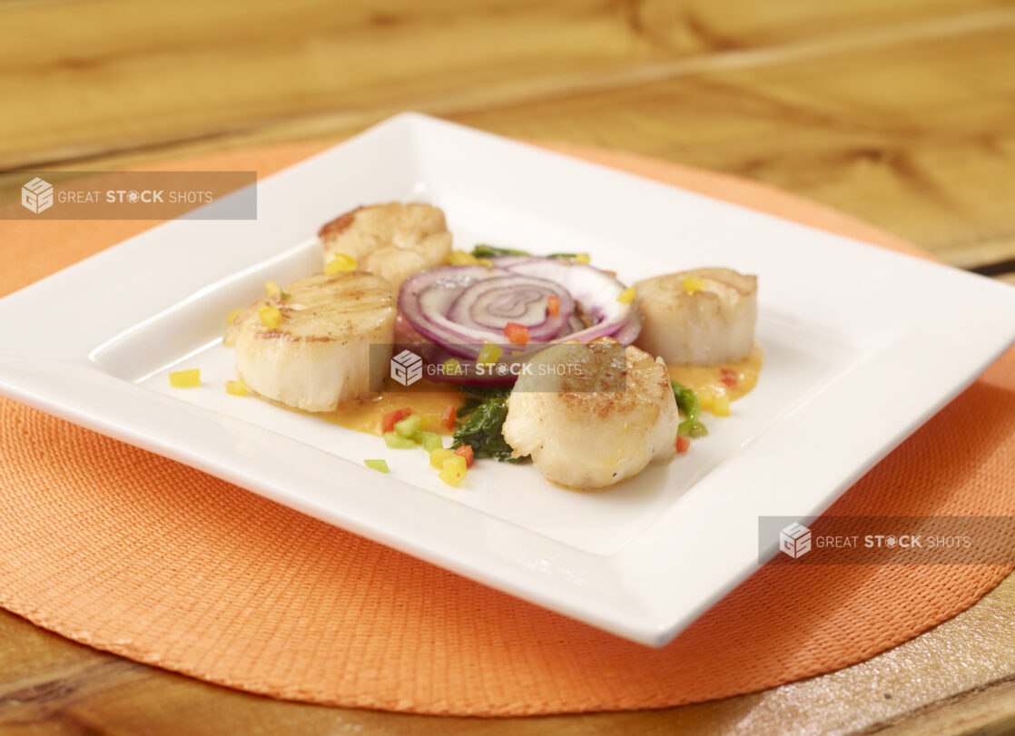 Pan Seared Scallops with Sauce on a Square White Dish with a Red Onion Slice on an Orange Placemat on a Wooden Table