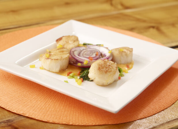 Pan Seared Scallops with Sauce on a Square White Dish with a Red Onion Slice on an Orange Placemat on a Wooden Table