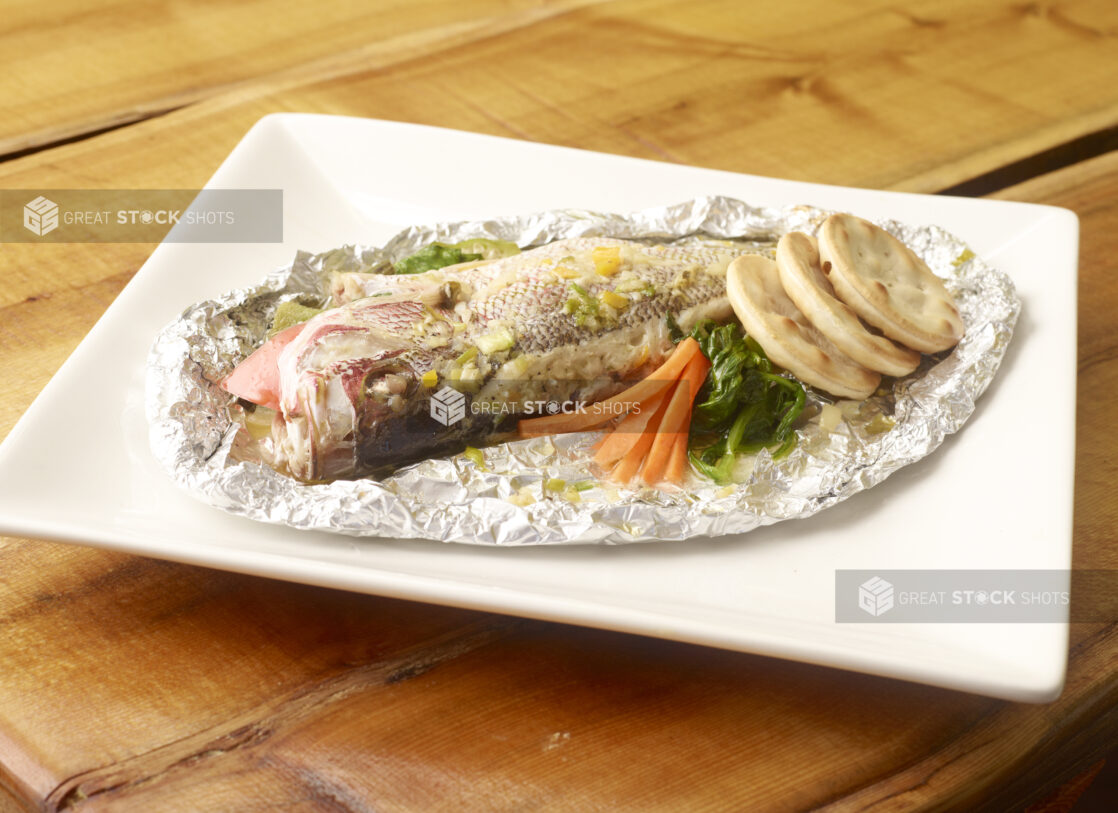 Whole Steamed Red Snapper Cooked in Foil Wrap with Cracker Biscuits on a Square White Platter on a Wooden Table