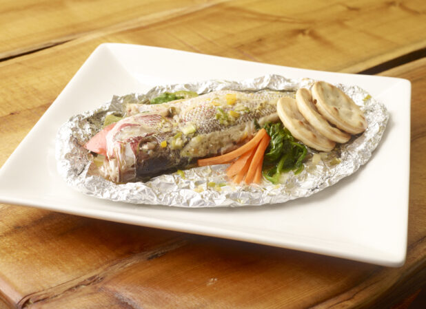 Whole Steamed Red Snapper Cooked in Foil Wrap with Cracker Biscuits on a Square White Platter on a Wooden Table