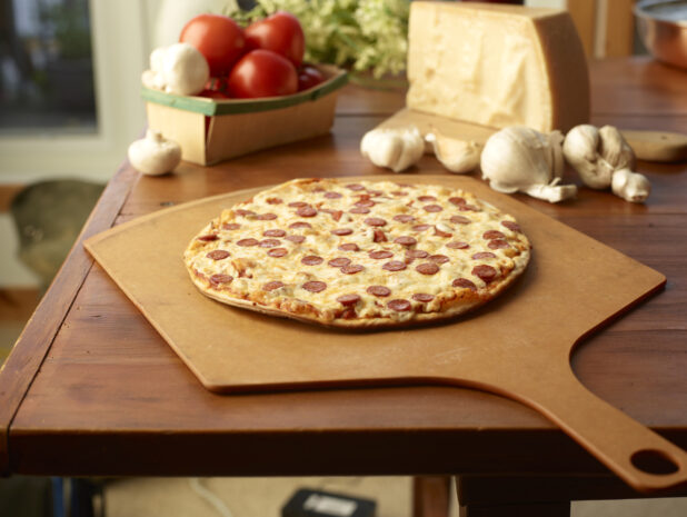Whole Pepperoni Pizza on a Wooden Pizza Peel, Surrounded by Fresh Pizza Ingredients on a Wooden Table in a Restaurant Interior