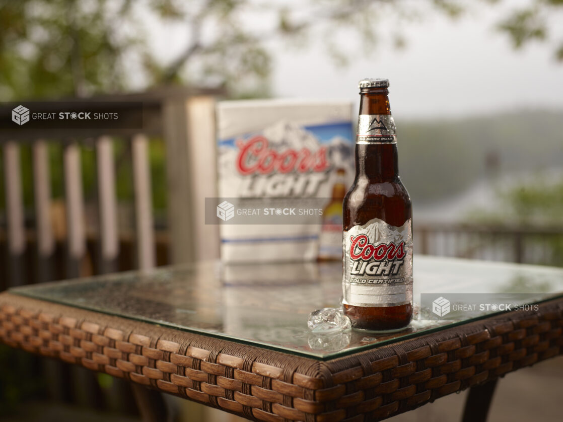 Brown Glass Bottle of Coors Light Beer on a Patio Table with a 6-Pack of Bottled Coors Light Beer in an Outdoor Setting