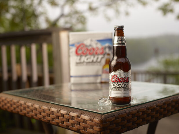 Brown Glass Bottle of Coors Light Beer on a Patio Table with a 6-Pack of Bottled Coors Light Beer in an Outdoor Setting