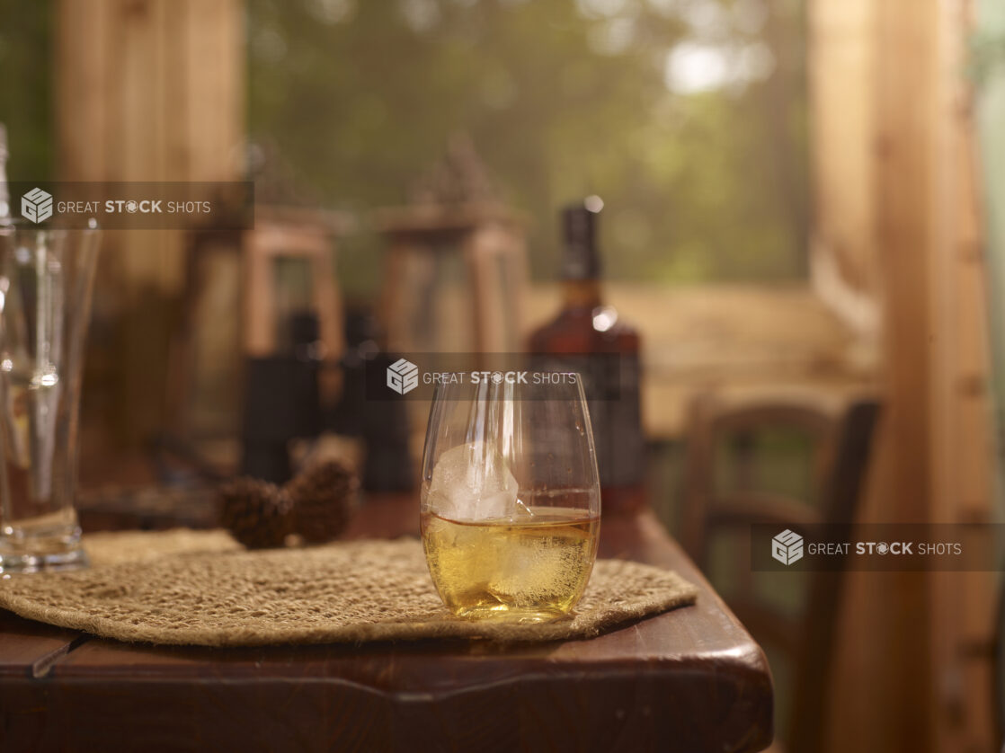 Stemless Glassware with Two Fingers of Jack Daniels Whiskey on the Rocks on a Woven Placemat and Wooden Table in an Indoor Setting