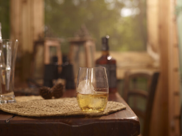 Stemless Glassware with Two Fingers of Jack Daniels Whiskey on the Rocks on a Woven Placemat and Wooden Table in an Indoor Setting