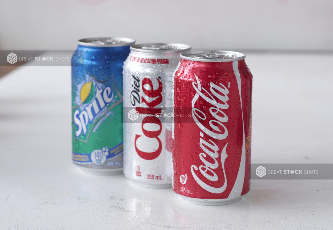 A Trio of Assorted Coca-Cola Beverage Soft Drink Cans on a White Surface in an Indoor Setting