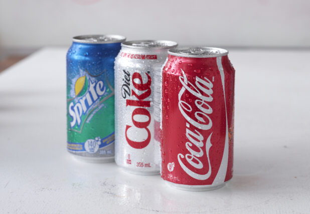 A Trio of Assorted Coca-Cola Beverage Soft Drink Cans on a White Surface in an Indoor Setting