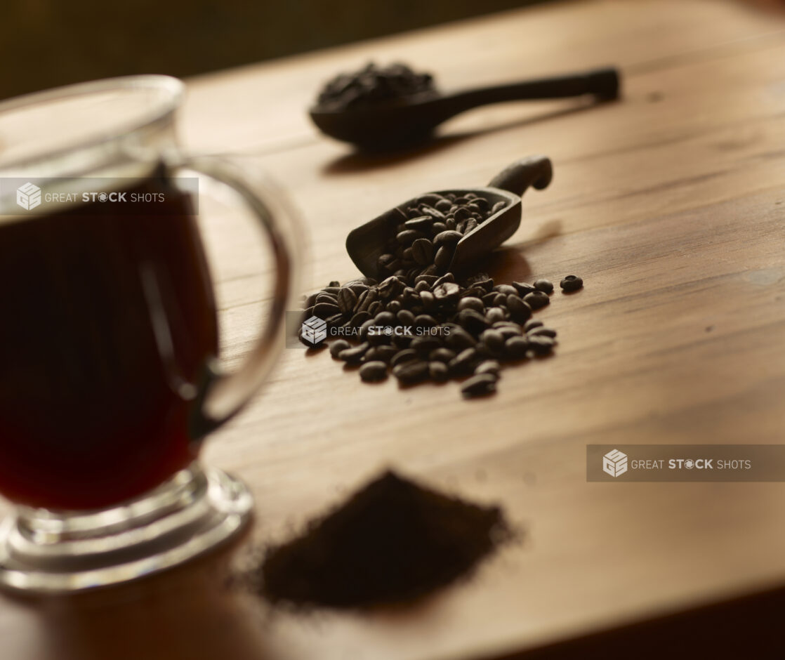 Whole Roasted Coffee Beans Spilling Out from a Wooden Scoop on a Wooden Surface with a Glass Mug of Coffee and Ground Coffee in an Indoor Setting