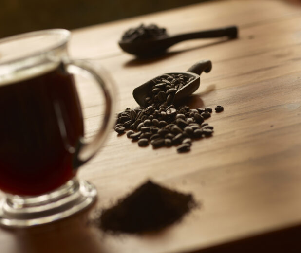 Whole Roasted Coffee Beans Spilling Out from a Wooden Scoop on a Wooden Surface with a Glass Mug of Coffee and Ground Coffee in an Indoor Setting