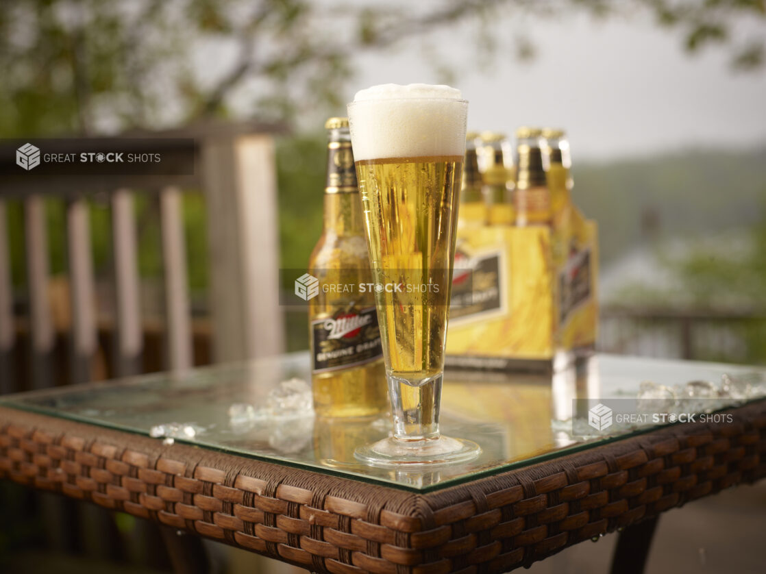 Half Pint Glass and Clear Glass Bottle of Miller Light Beer on a Patio Table with a 6-Pack of Bottled Miller Light Beer in an Outdoor Setting