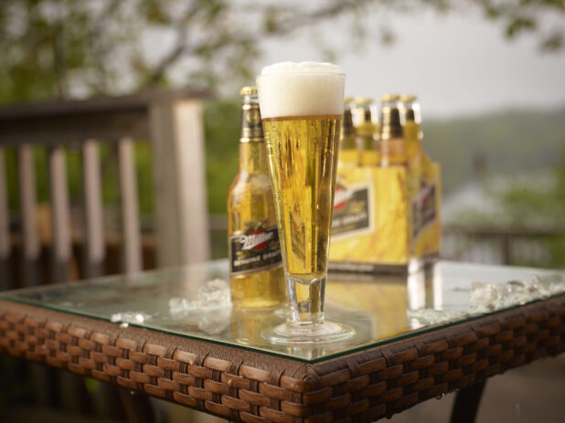 Half Pint Glass and Clear Glass Bottle of Miller Light Beer on a Patio Table with a 6-Pack of Bottled Miller Light Beer in an Outdoor Setting