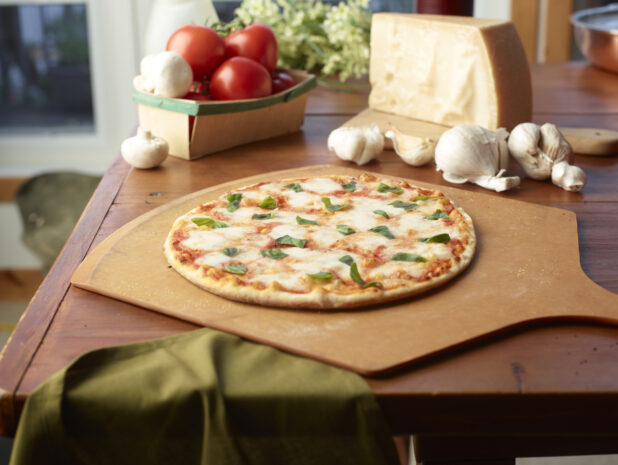 Whole Margherita Pizza on a Wooden Pizza Peel, Surrounded by Fresh Pizza Ingredients on a Wooden Table in a Restaurant Interior