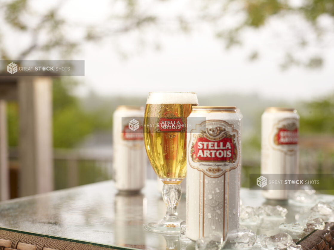 Stella Artois Tall Boy Cans and a Half-Pint Glass on a Patio Table with Ice Cubes in an Outdoor Setting