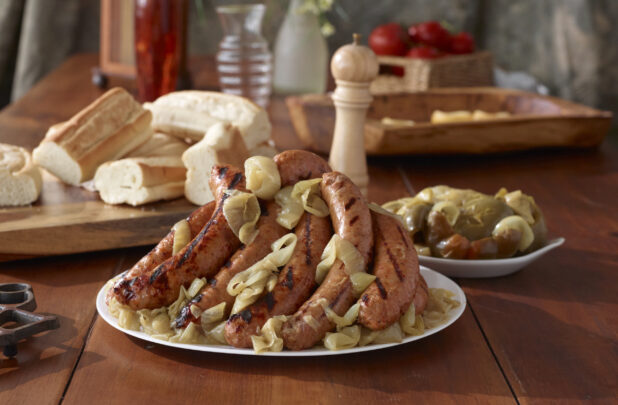 Platter of Grilled Sausage Links and Roasted Onions with a Side Dish of Cooked Peppers and Sliced Baguettes on a Wooden Table Indoors