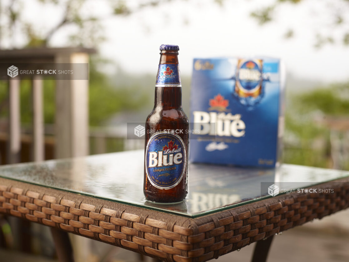 Brown Glass Bottle of Labatt Blue Beer on a Patio Table with a 6-Pack of Bottled Labatt Blue Beer in an Outdoor Setting