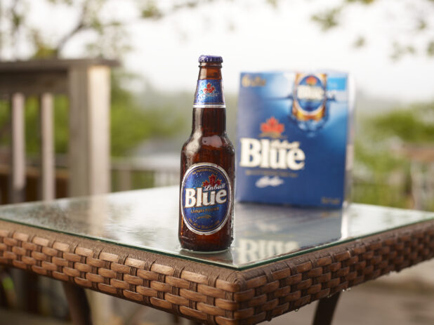 Brown Glass Bottle of Labatt Blue Beer on a Patio Table with a 6-Pack of Bottled Labatt Blue Beer in an Outdoor Setting
