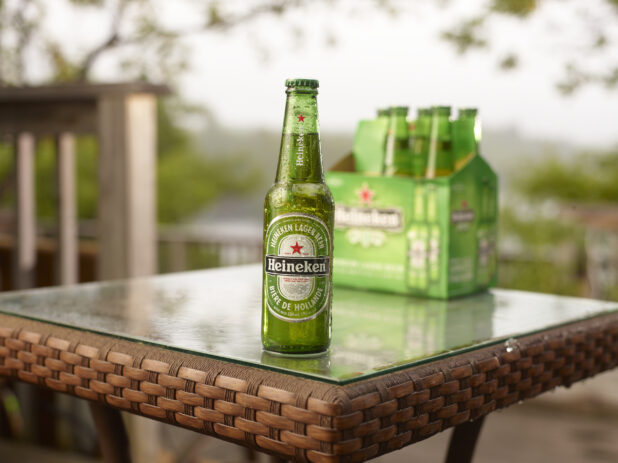 Green Glass Bottle of Heineken Beer on a Patio Table with a 6-Pack of Bottled Heineken Beer in an Outdoor Setting