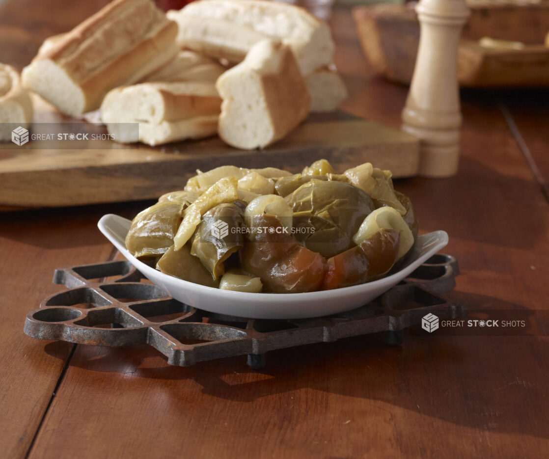 Side Dish of Oven Roasted Green and Red Peppers and Onions in a White Ceramic Dish on a Cast Iron Trivet and Wooden Table with Sliced Baguettes in an Indoor Setting