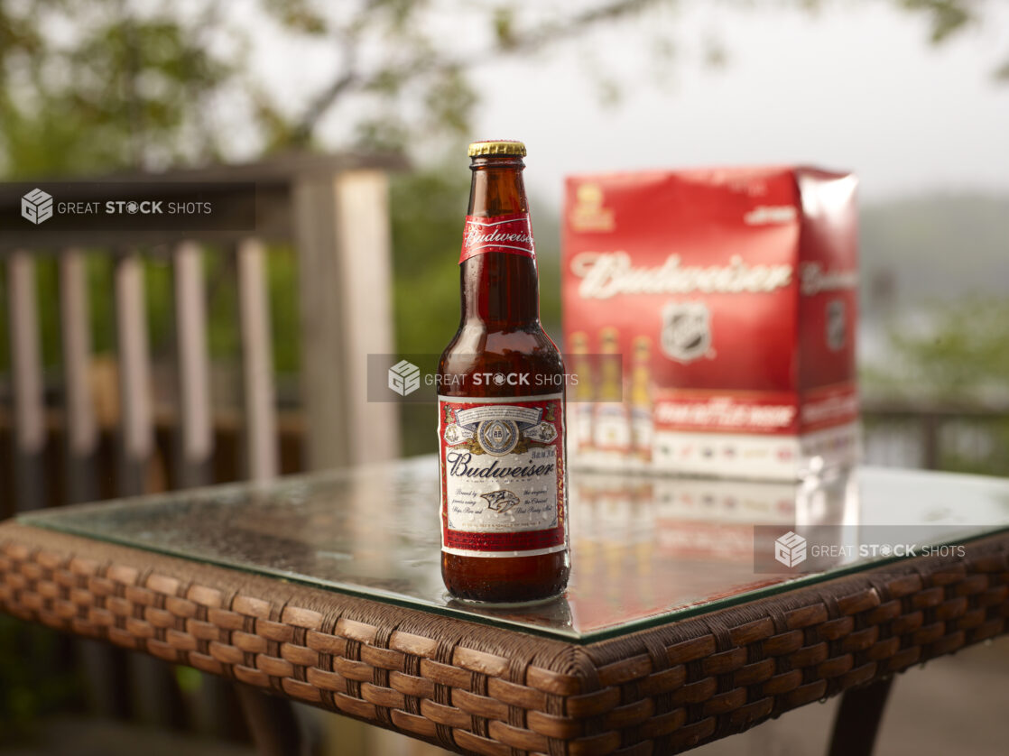 Brown Glass Bottle of Budweiser Beer on a Patio Table with a 6-Pack of Bottled Budweiser Beer in an Outdoor Setting