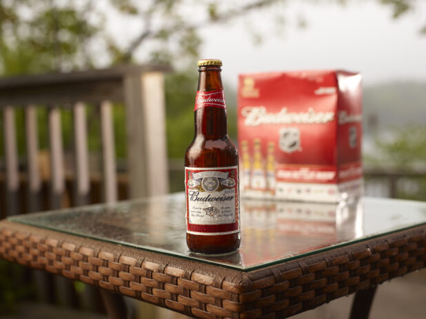 Brown Glass Bottle of Budweiser Beer on a Patio Table with a 6-Pack of Bottled Budweiser Beer in an Outdoor Setting